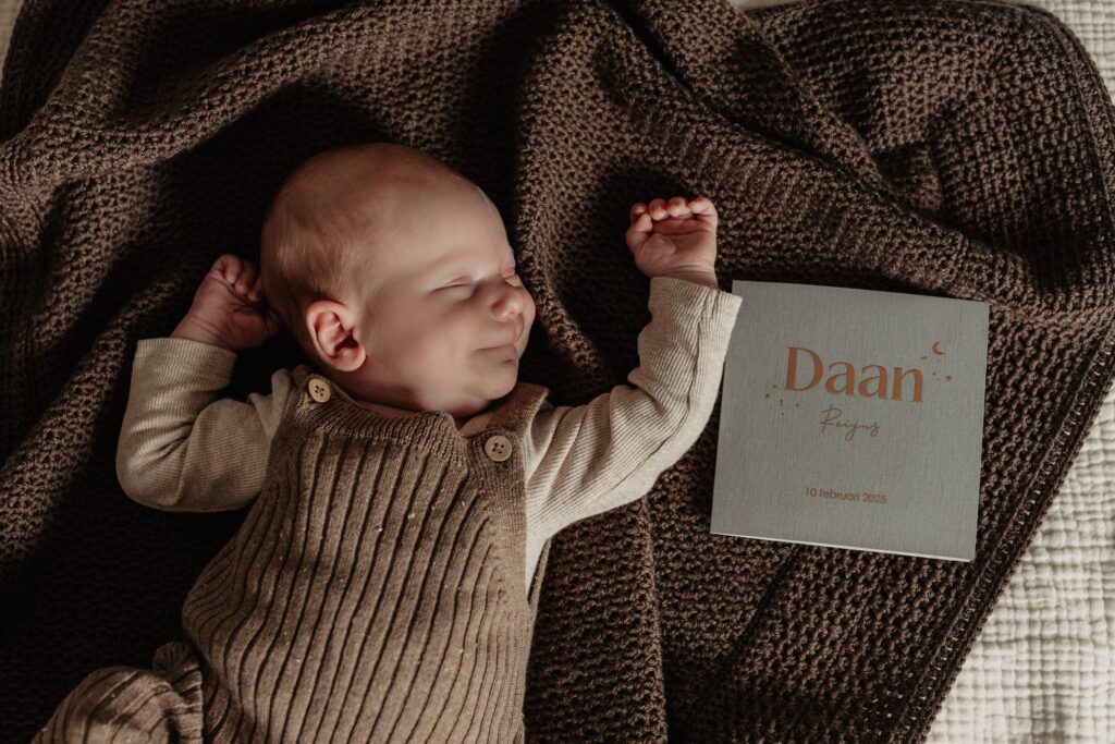 Pasgeboren baby ligt op een bruin gebreid dekentje met geboortekaartje tijdens een newbornshoot aan huis.